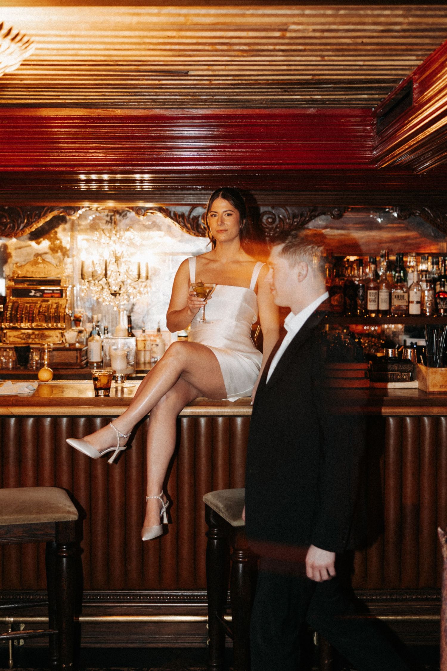 Elegant couple at a luxurious bar with chandelier lighting and vintage decor in warm ambient setting.