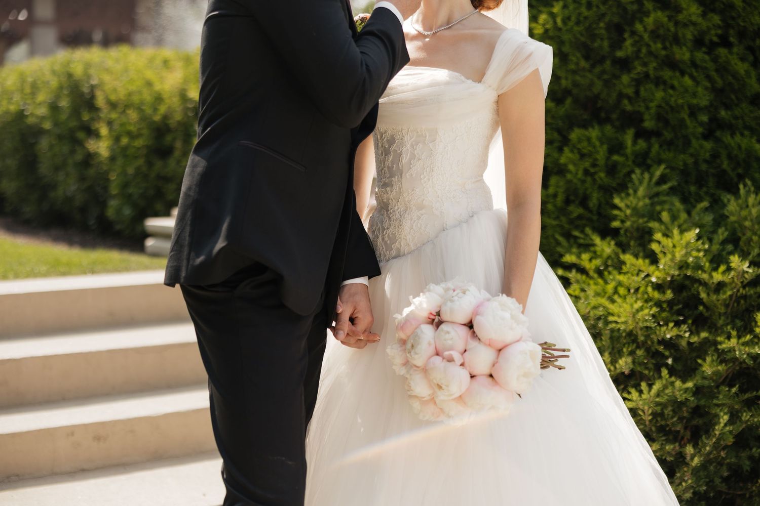 Bride in white ball gown holding pink peony bouquet stands with groom in black suit on outdoor steps.