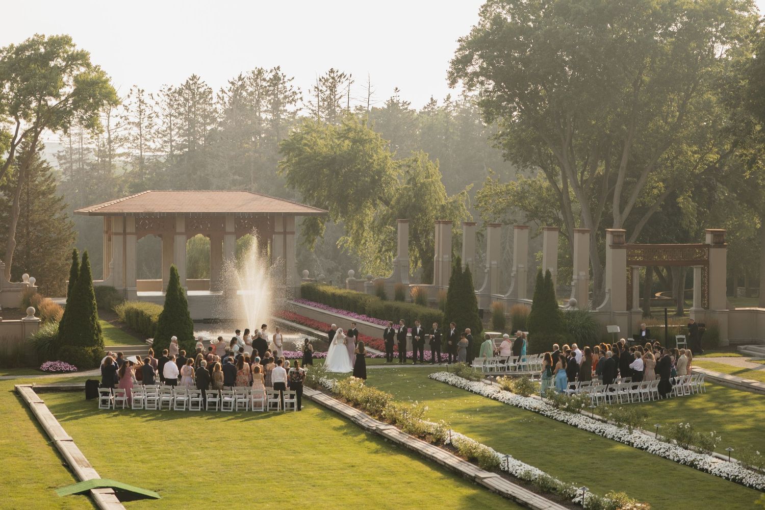 Elegant outdoor wedding ceremony in a lush garden with a gazebo fountain, manicured hedges, and guests seated on white chairs.