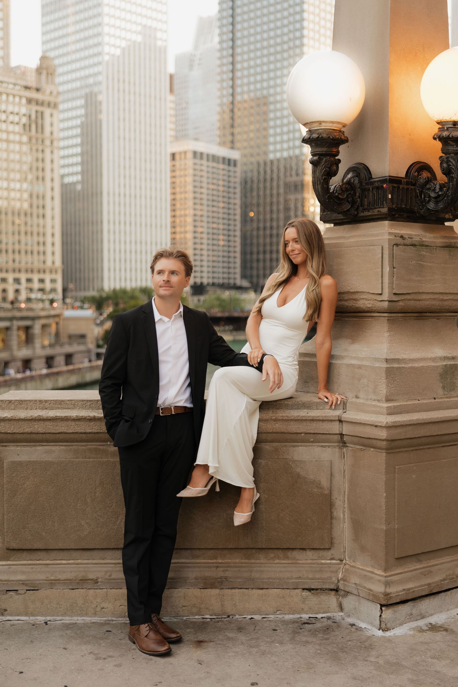 Elegant couple poses by ornate Chicago bridge lamp, man in black suit, woman in white dress, city skyline backdrop.