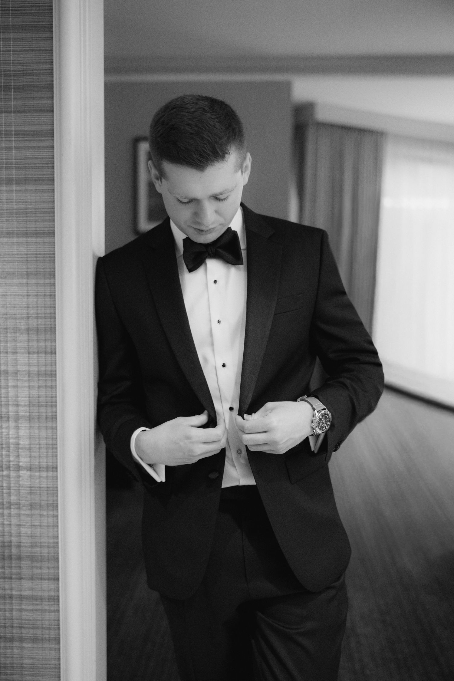 Black and white photo of a groom in a tuxedo and bow tie buttoning his jacket before his wedding ceremony.