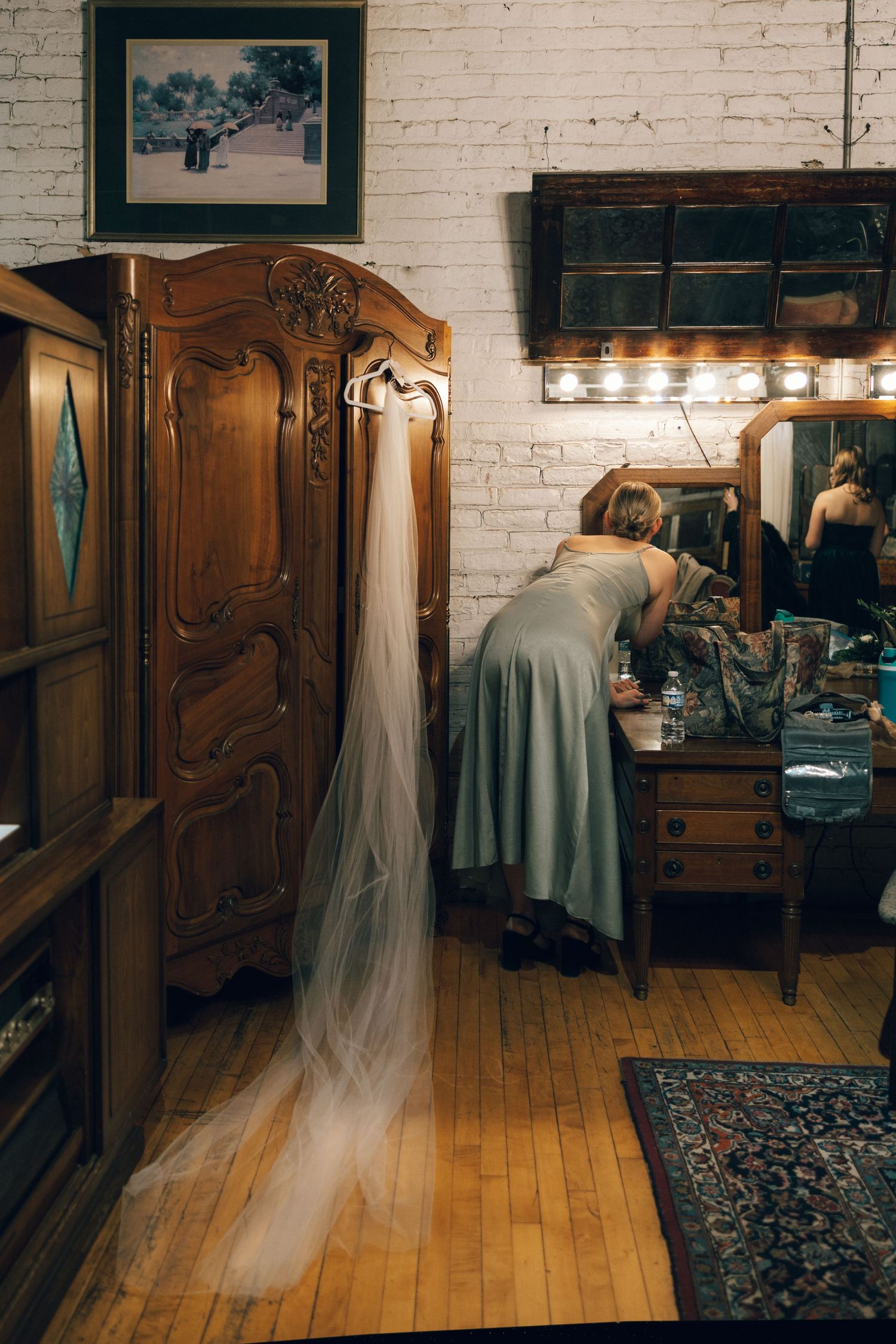 A wedding dress hangs from an ornate wooden armoire while someone prepares at a vintage vanity in a rustic room.