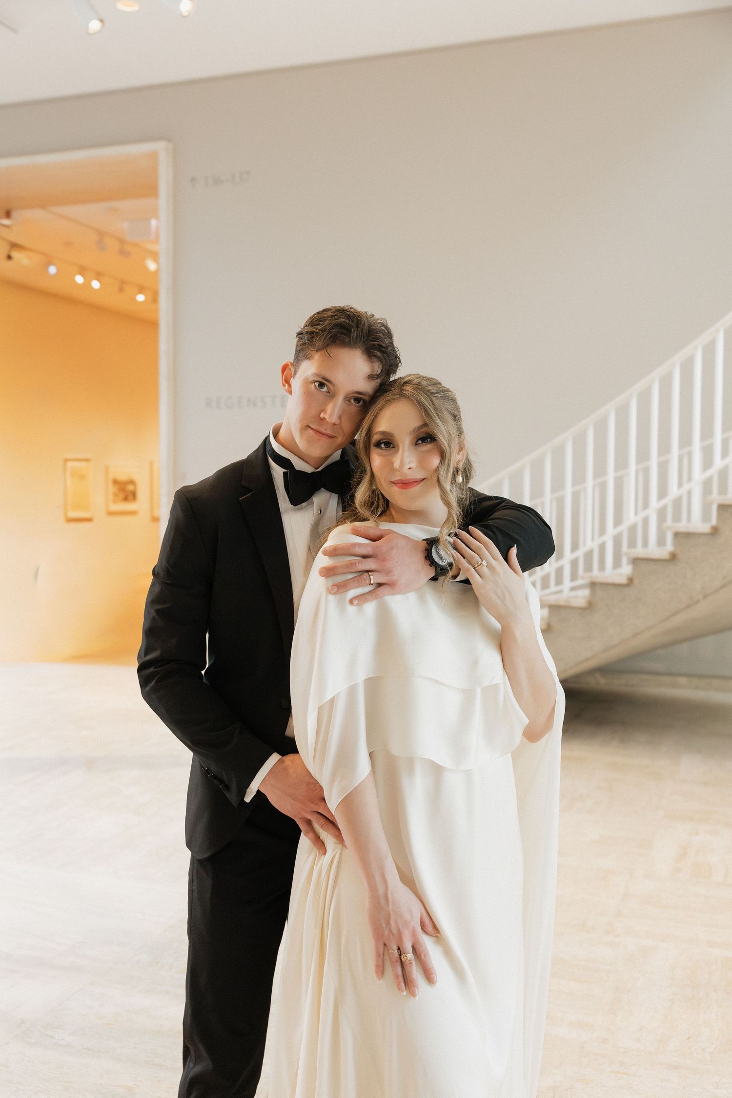 A wedding couple poses together in an elegant indoor setting with white stairs in the background.