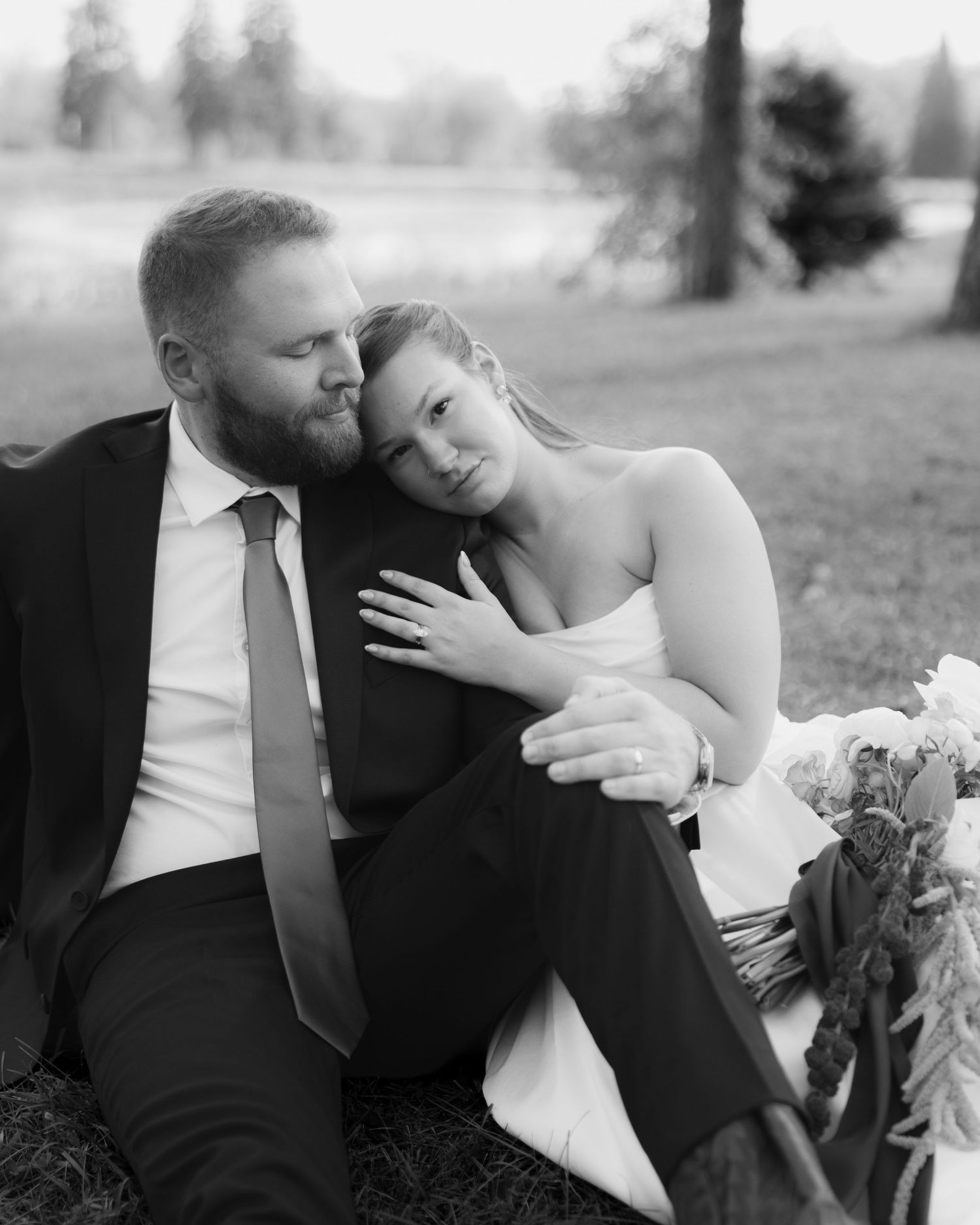 Black and white photo of a bride leaning her head on a groom's shoulder while seated on the grass with a bouquet nearby.