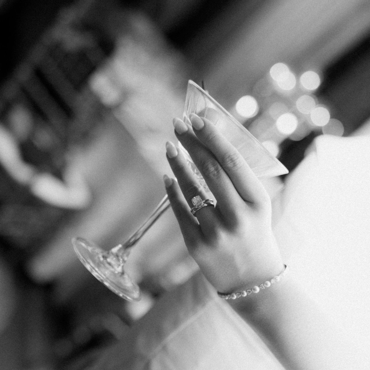 Black and white close-up of a woman's hand with a ring and pearl bracelet holding a martini glass at an elegant evening event.