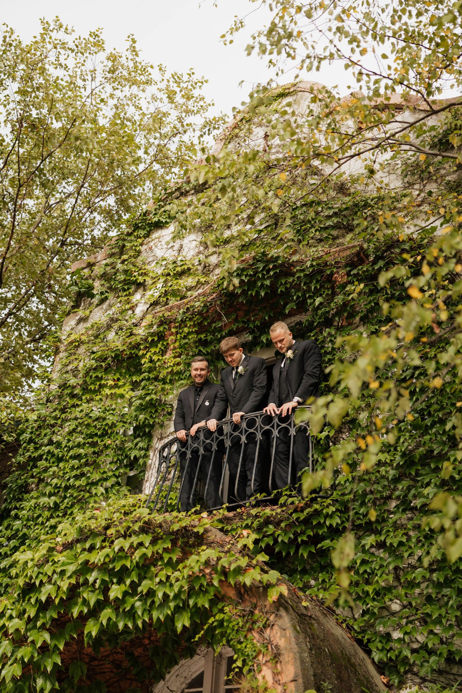 Three groomsmen in dark suits lean on a wrought iron balcony railing of an ivy-covered stone building at an autumn wedding.