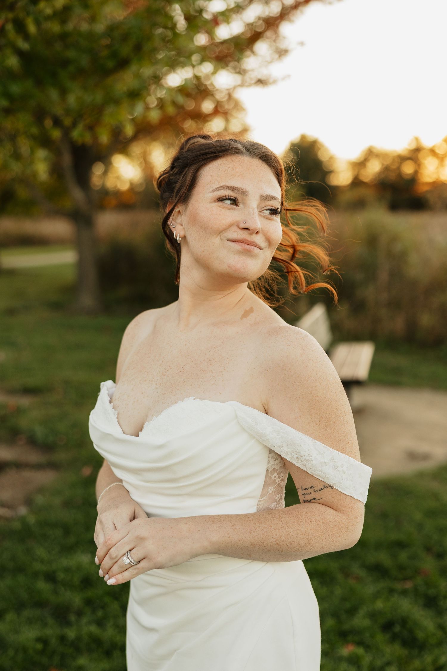 Smiling bride in an off-the-shoulder white satin wedding dress poses outdoors in a park at golden hour with trees and a bench behind her.