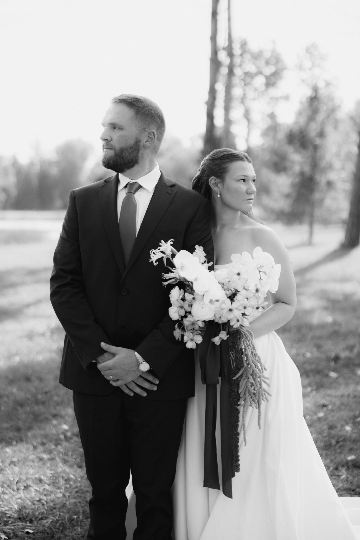 Black and white wedding portrait of a bearded groom in a suit and a bride in a white gown holding a bouquet with trailing ribbons outdoors.