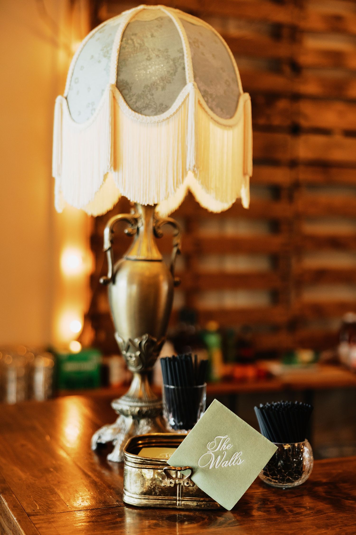 Vintage brass table lamp with a cream fringed shade on a wooden bar beside a small card reading The Walls and a jar of black straws.