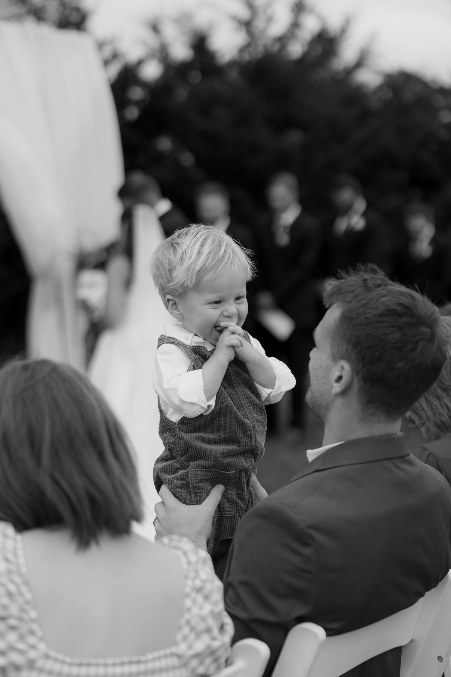 Black and white photo of a smiling toddler in overalls held up by a guest during an outdoor wedding ceremony.