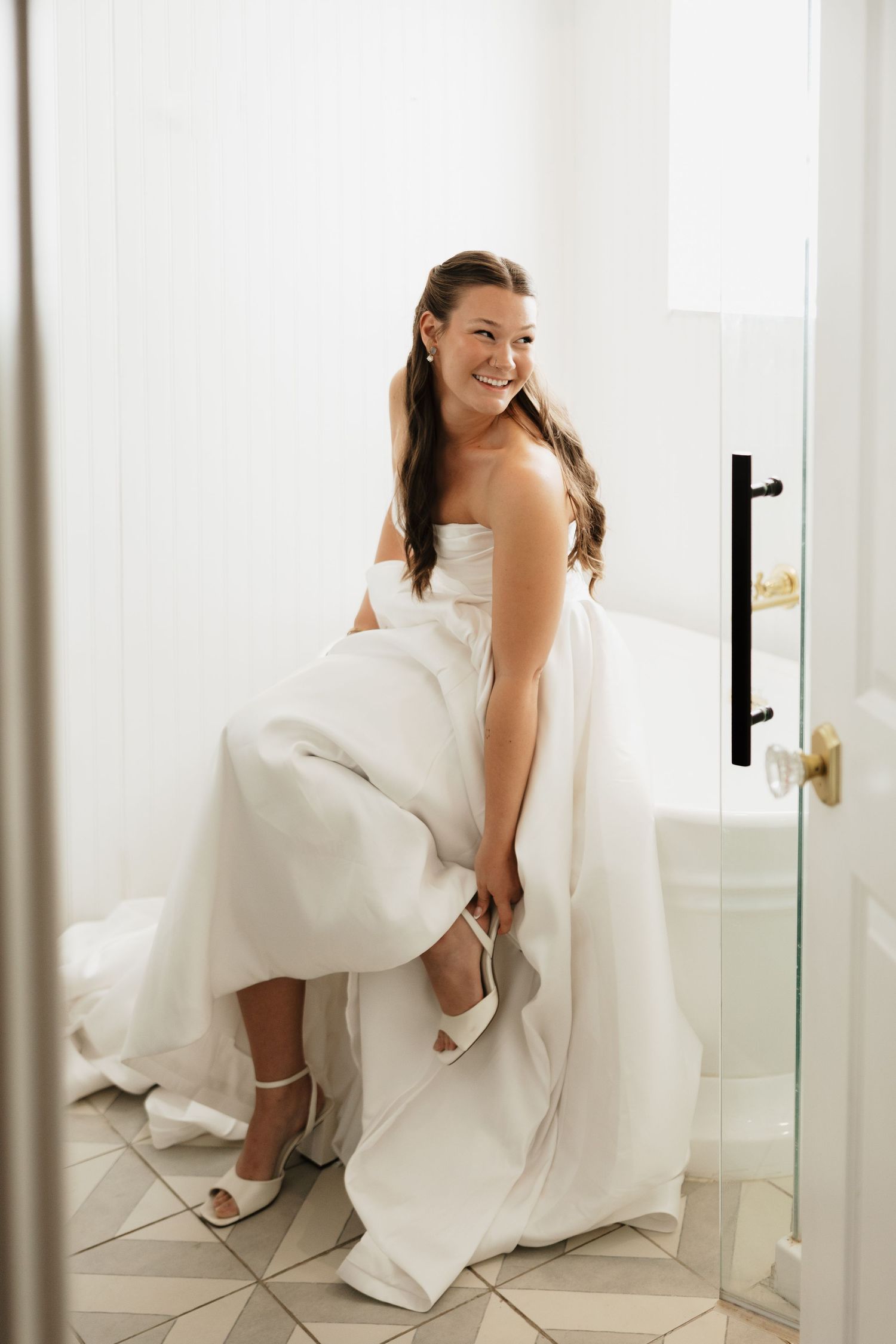 Smiling bride in a strapless white wedding gown sits on a bathtub edge while putting on her white strappy wedding heels.