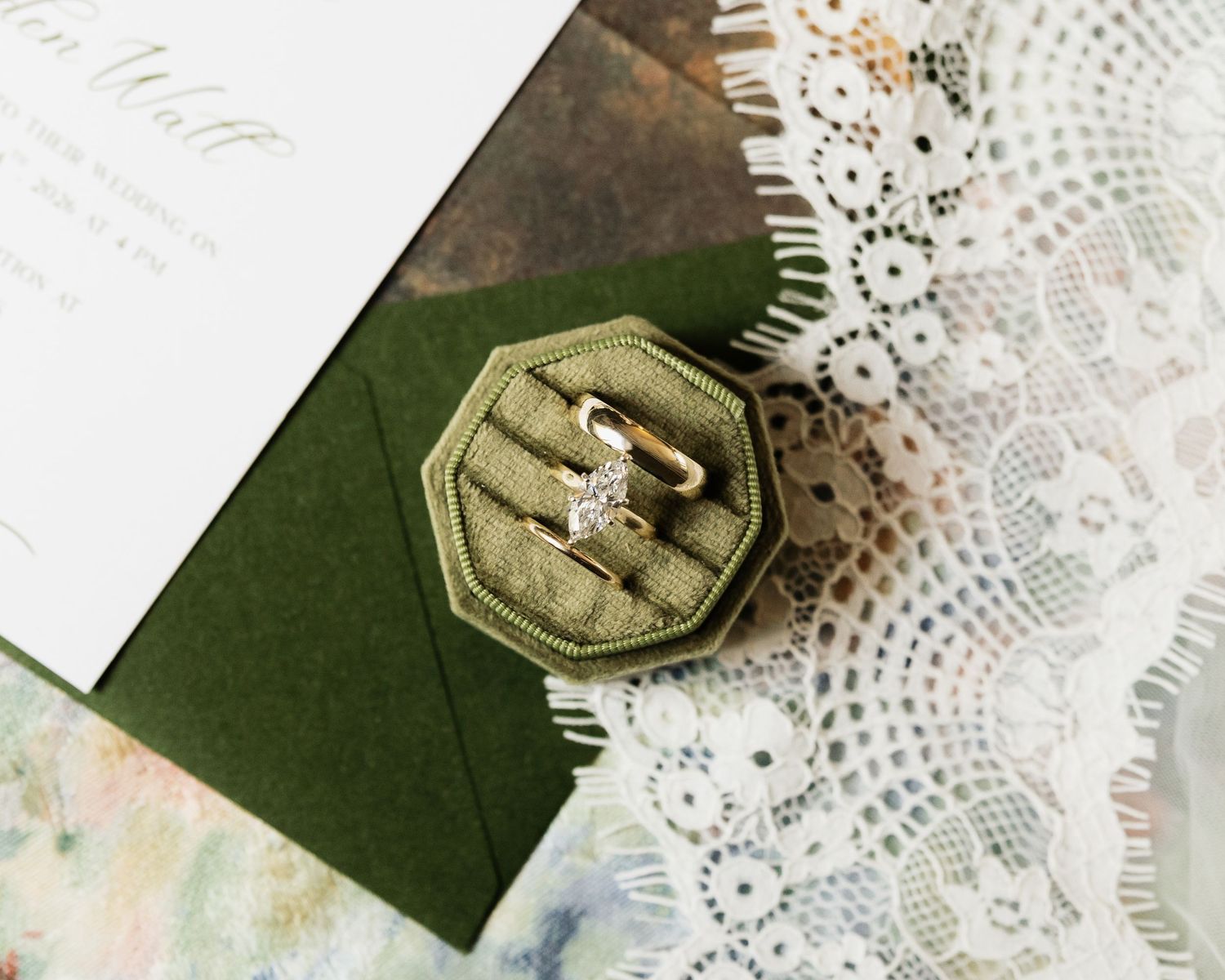 Diamond engagement ring and wedding band set in an open olive green velvet octagonal ring box beside white lace and a wedding invitation.