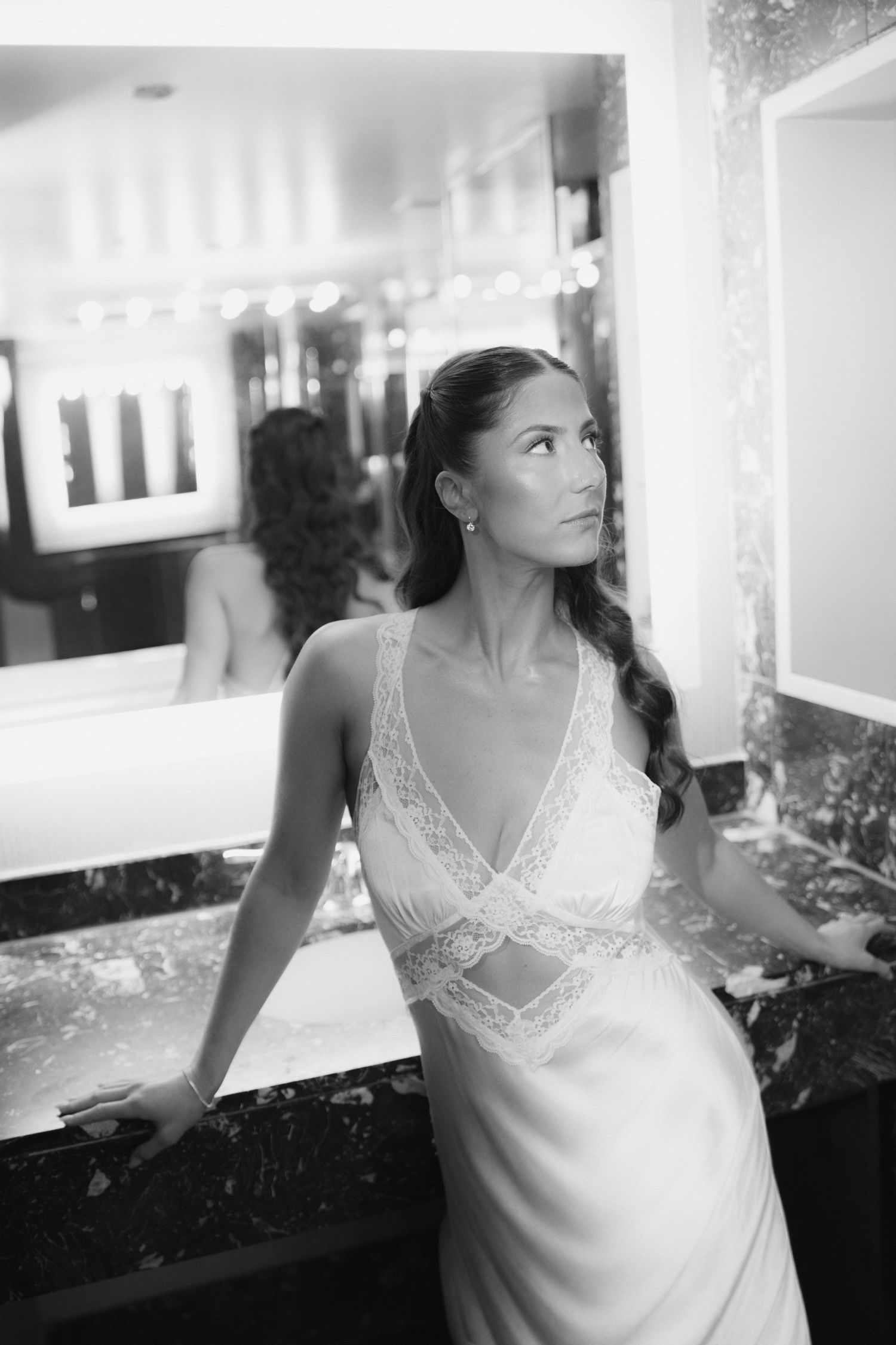 Black and white photo of a bride in a lace wedding gown standing by a marble vanity in an elegant bathroom.