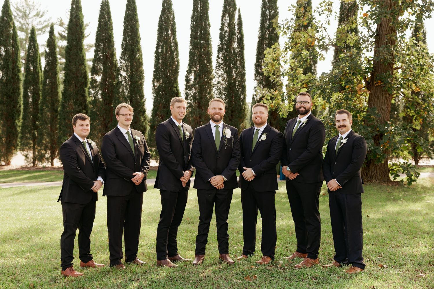 Seven groomsmen in dark suits and boutonnieres stand side by side on a sunny lawn in front of tall evergreen trees.
