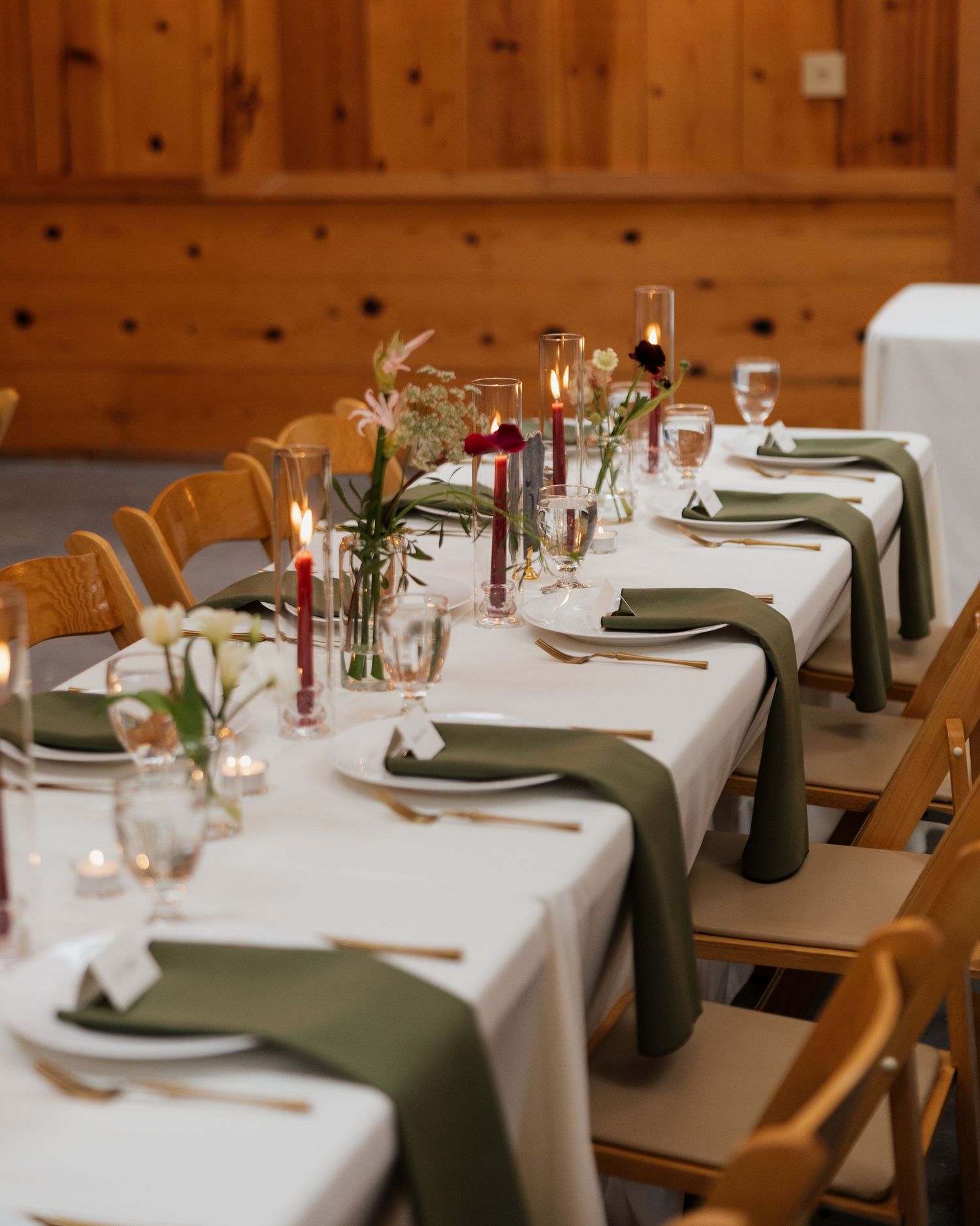 Long wedding reception table with olive green napkins, red taper candles, and floral centerpieces in a rustic wooden venue.