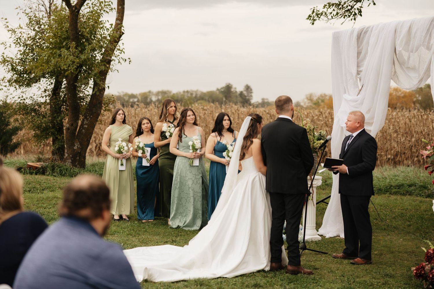 Outdoor fall wedding ceremony with a bride and groom facing an officiant as bridesmaids in green and teal dresses stand nearby.