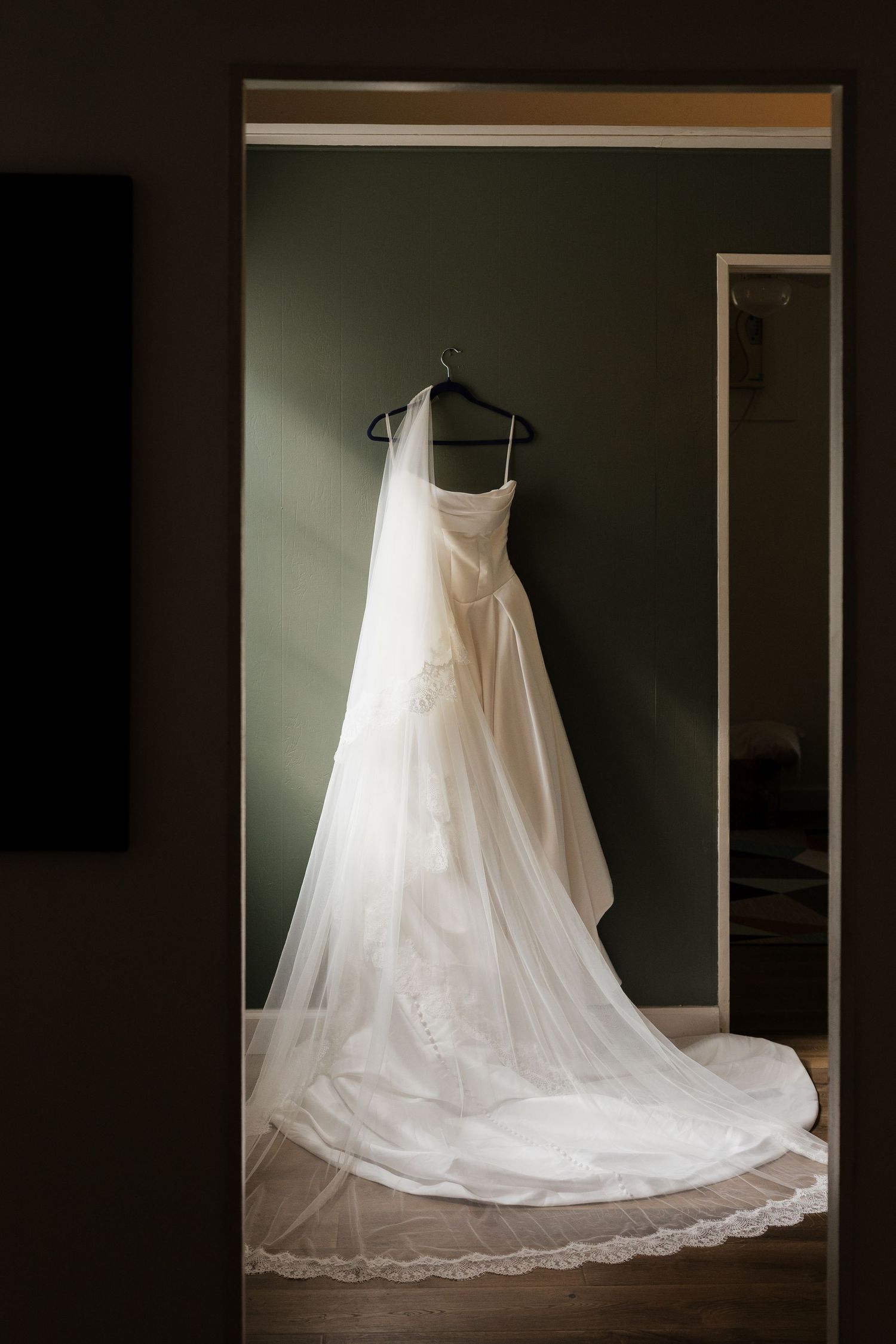 White strapless wedding gown with a long flowing veil hanging on a hanger in a doorway against a green wall.