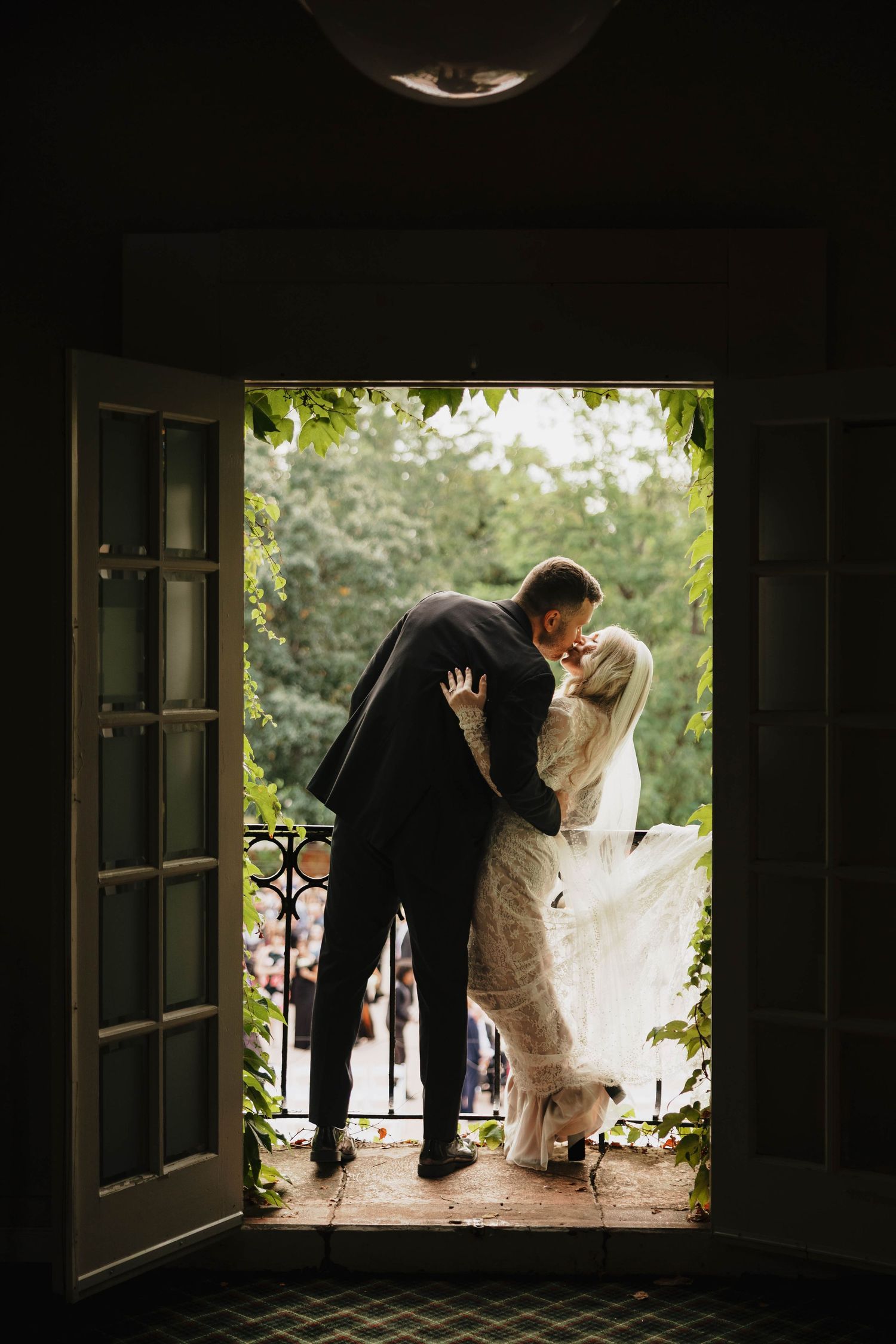Bride and groom share a kiss on a stone balcony framed by open French doors, with guests and green trees visible below.