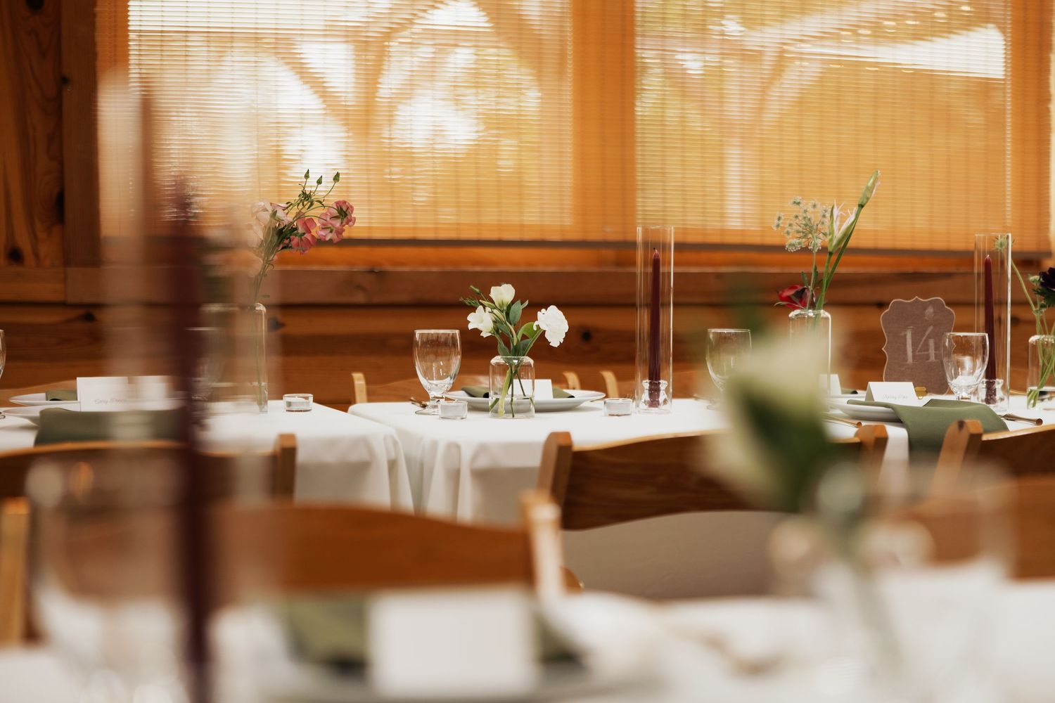 Elegant restaurant dining room with white-clothed tables, fresh flower centerpieces, wine glasses and wooden chairs by sunlit windows.