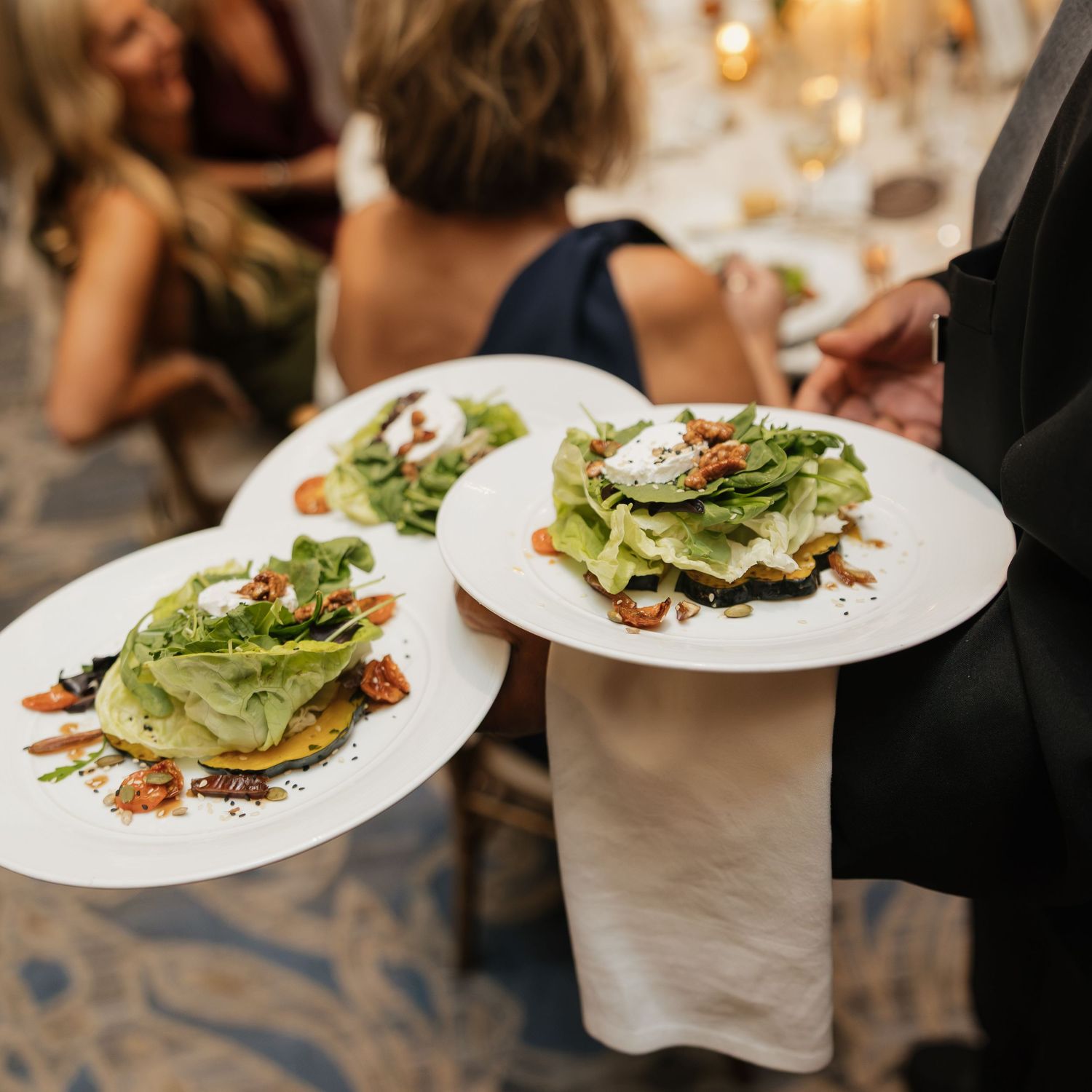 Server in a black jacket carries two white plates of fresh green salad topped with dressing and garnish at an elegant candlelit dinner.