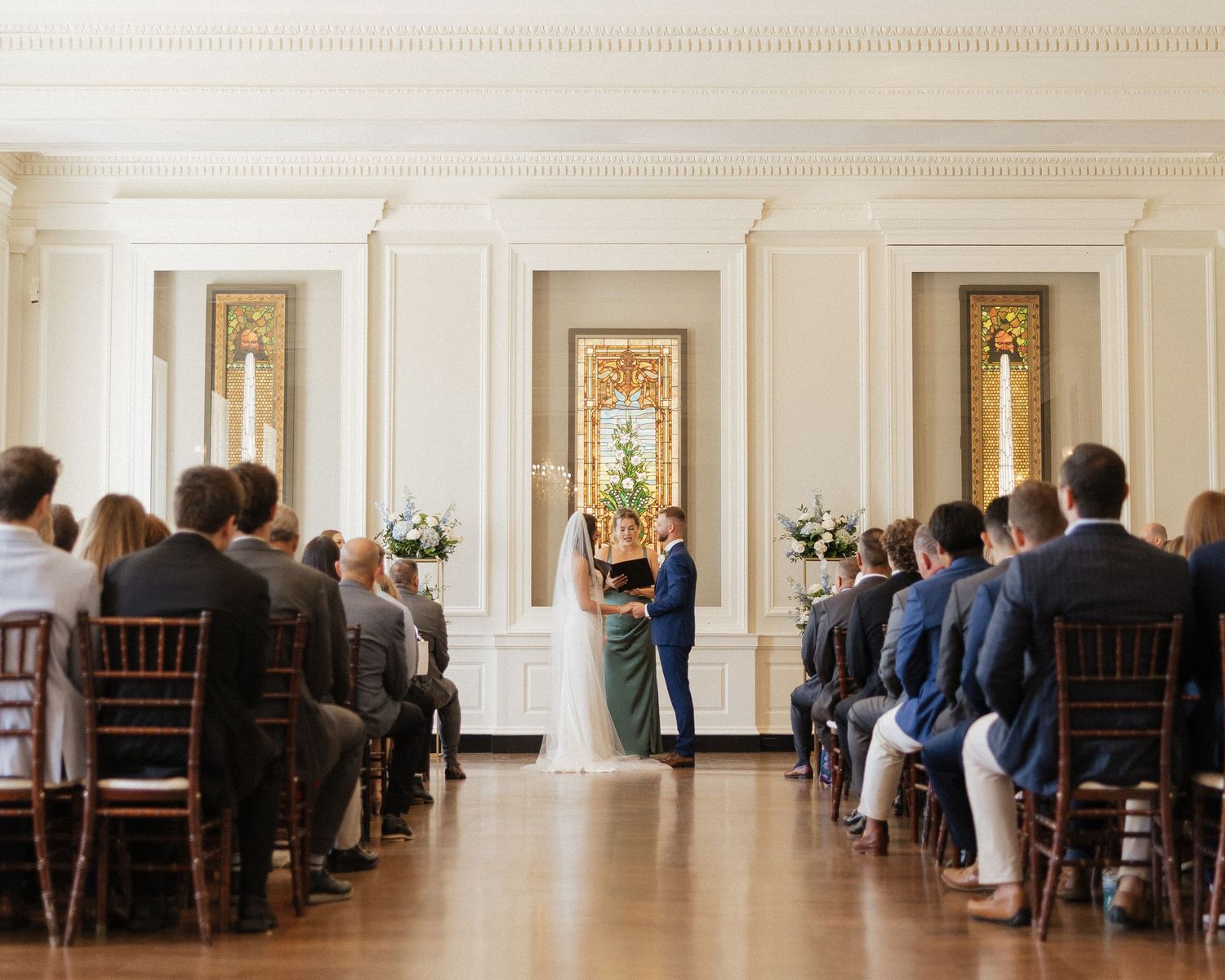 Panoramic view of a wedding ceremony in an elegant white room with dark wooden chairs and guests seated in rows.