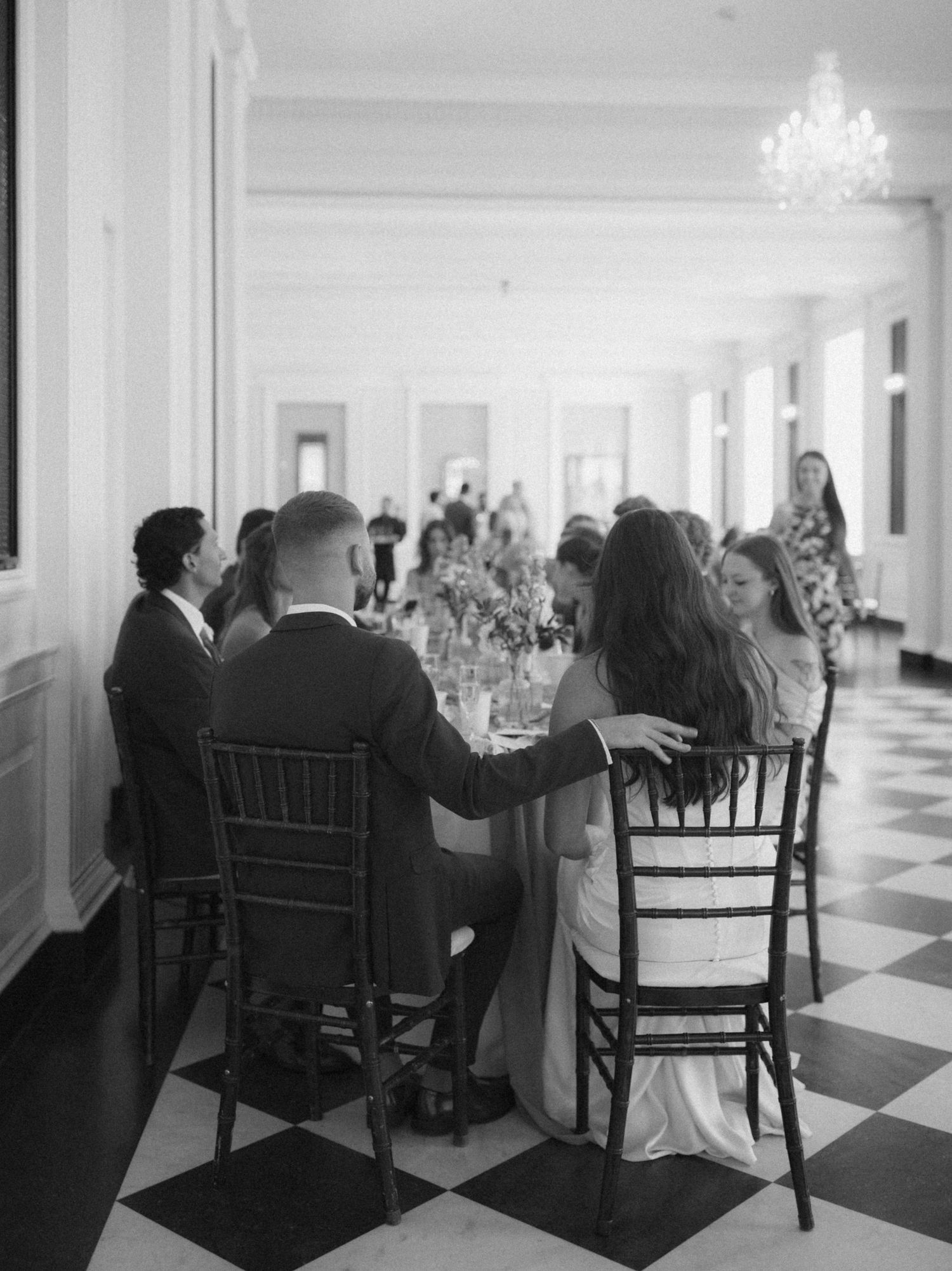Vintage black and white photograph of people sitting in rocking chairs along a long hallway with checkerboard floors.