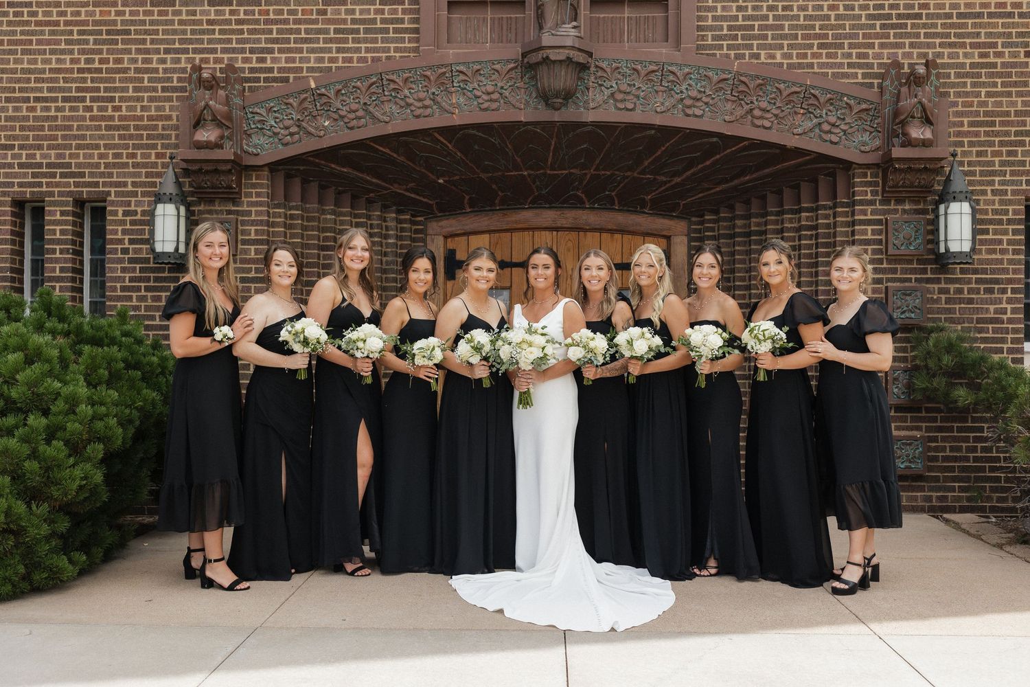 Wedding party in black dresses poses together against a brick building exterior.