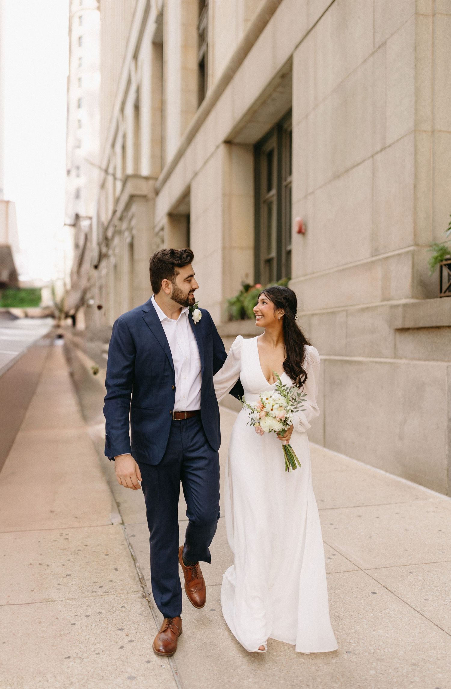A couple in wedding attire walking together along a city sidewalk while holding a white floral bouquet.