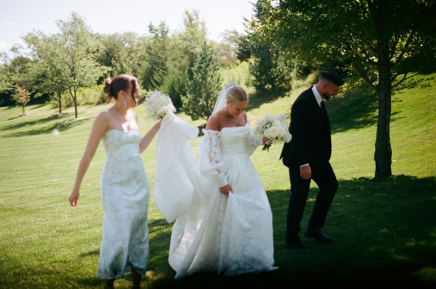 Three people in wedding attire walk together on a grassy hillside on a sunny day.