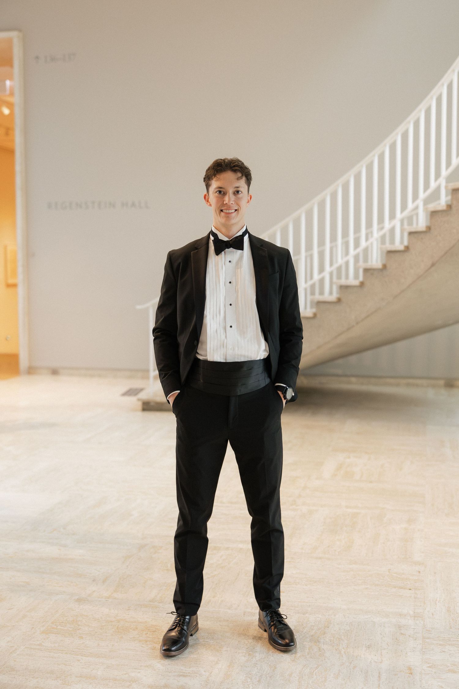 Young ring bearer in black tuxedo poses in elegant venue with white staircase.