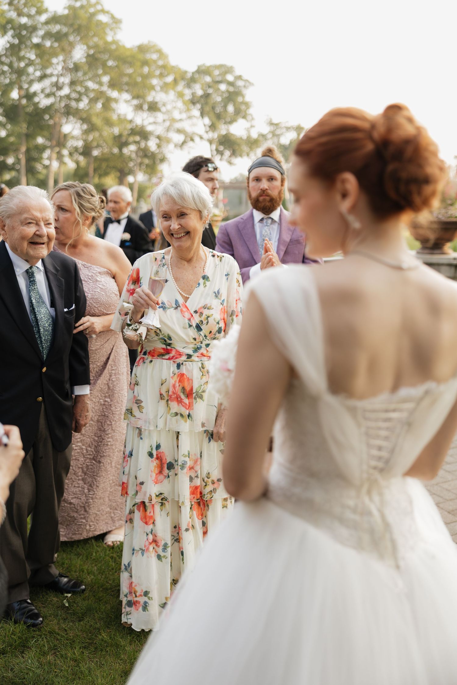 A red-haired bride in a white corset gown greets smiling wedding guests outdoors at an elegant garden celebration.