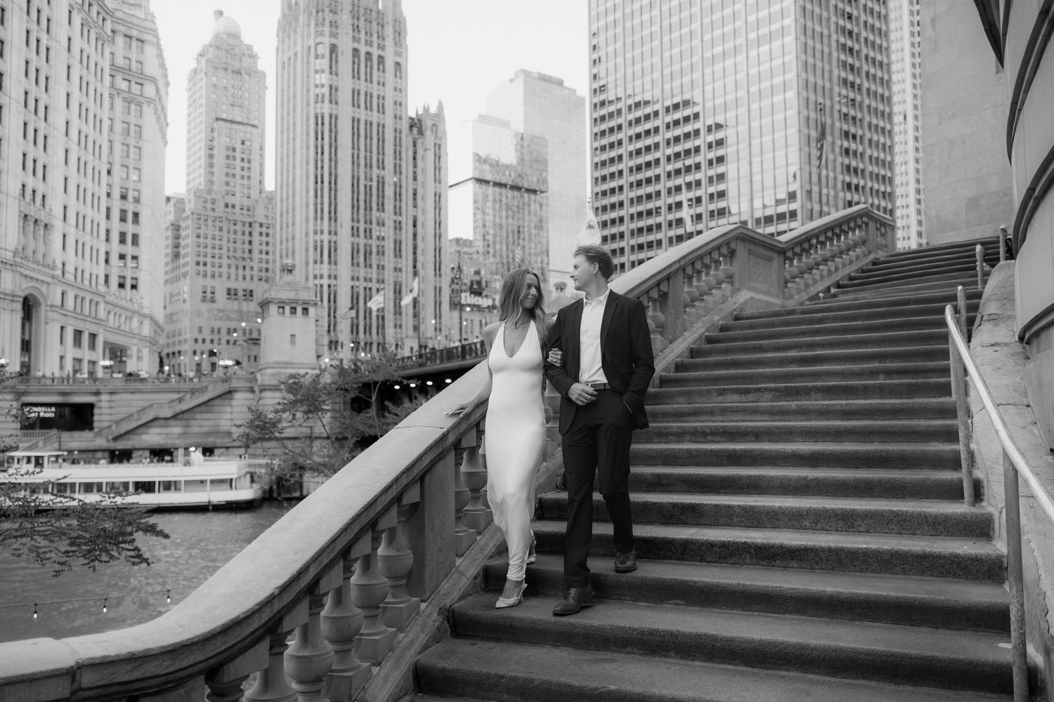 Black and white photo of a couple holding hands on grand stairs by the Chicago River with city skyline backdrop.