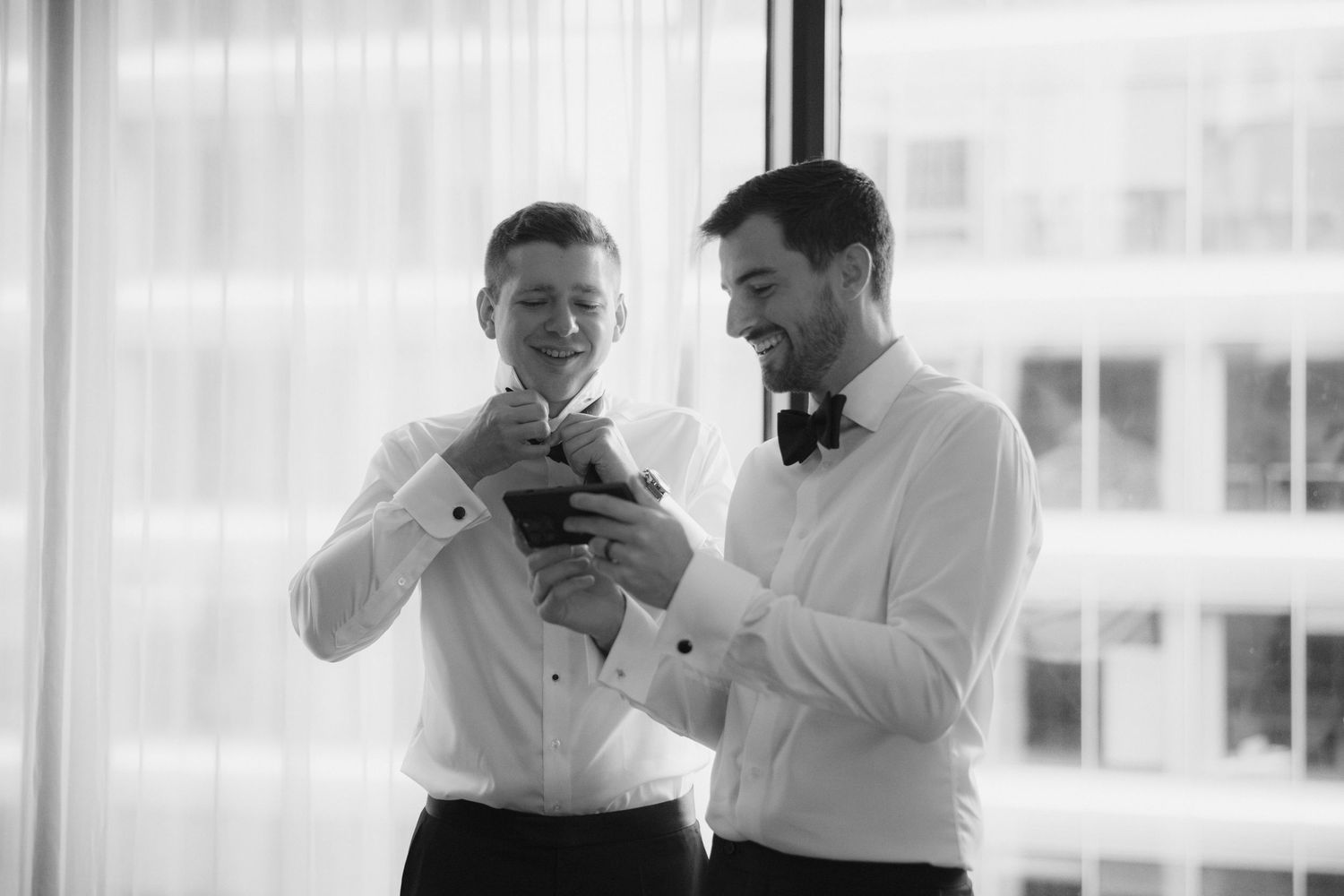 Two grooms in white shirts and bow ties share a joyful moment while looking at a gift before their wedding ceremony.
