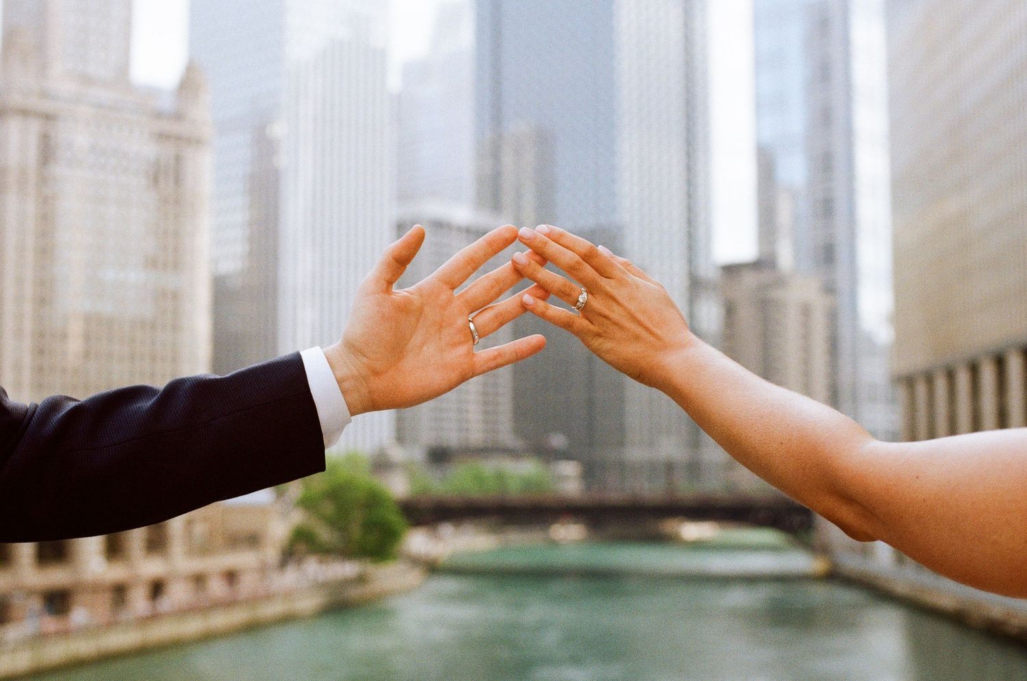 Couple touching fingertips showing wedding rings with Chicago River and city skyline in background.
