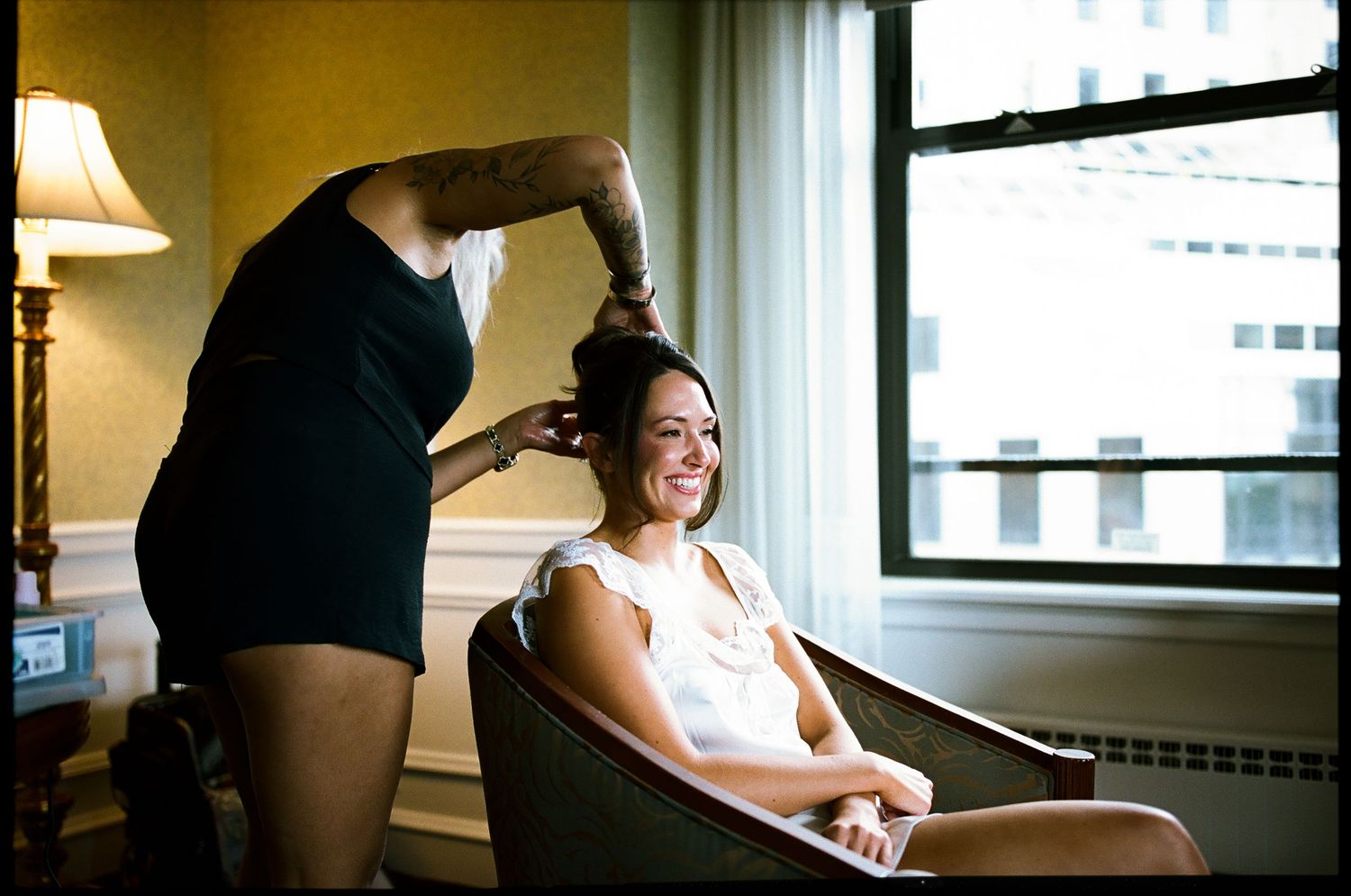 A smiling bride gets her hair styled by a hairdresser while seated in a chair near a bright window.