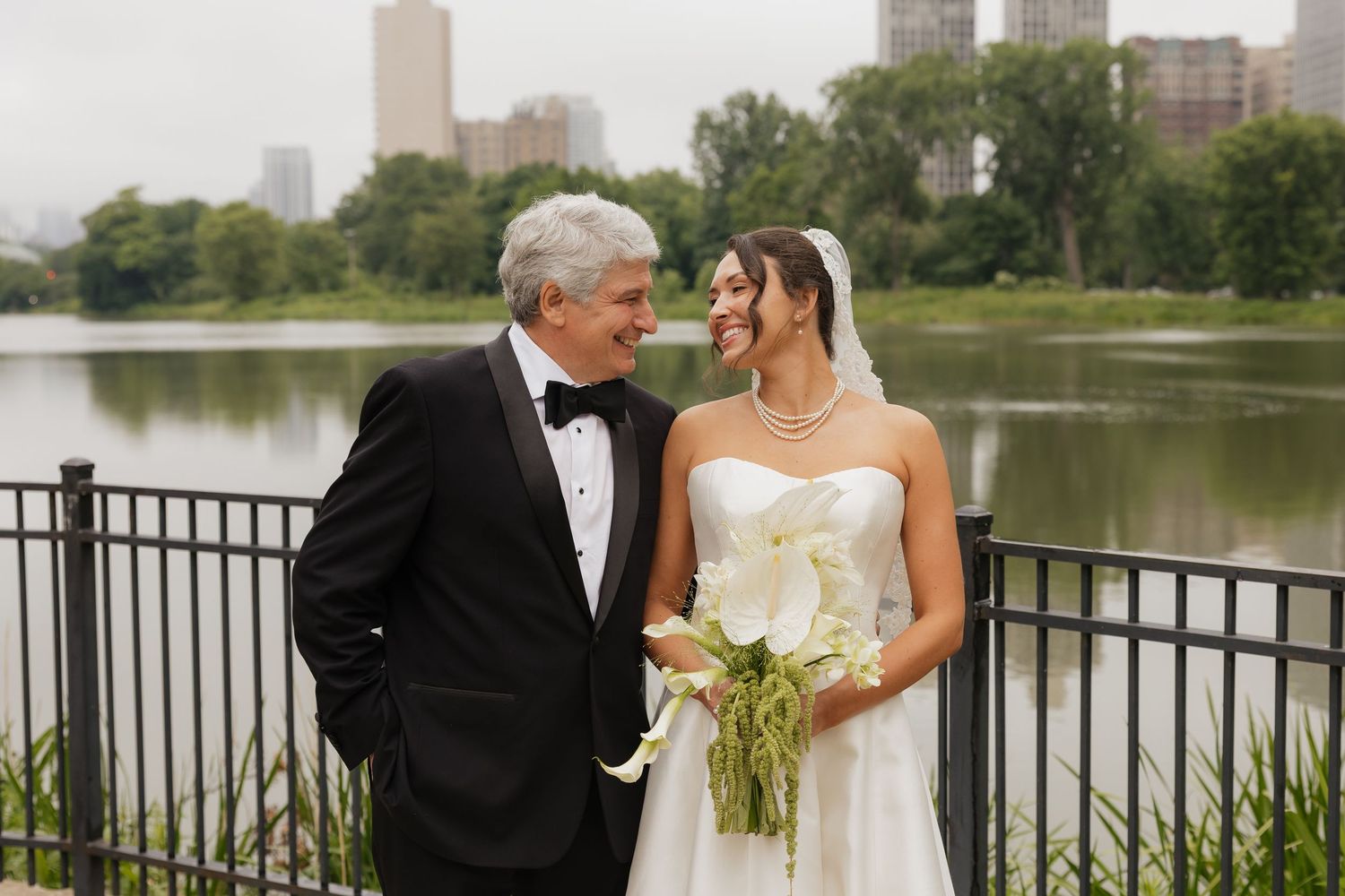 Bride and groom smiling at each other near a lakeside railing, bride holding white calla lily bouquet, city skyline in background.