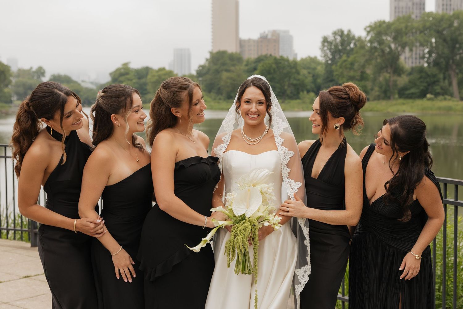 Bride in white gown with veil holds bouquet surrounded by bridesmaids in black dresses outdoors near a lake.