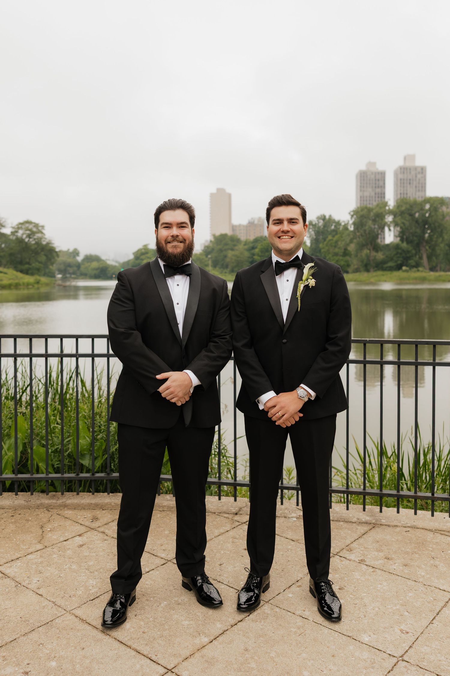 Two men wearing black tuxedos with bow ties smile outdoors by a serene lake with city buildings behind.