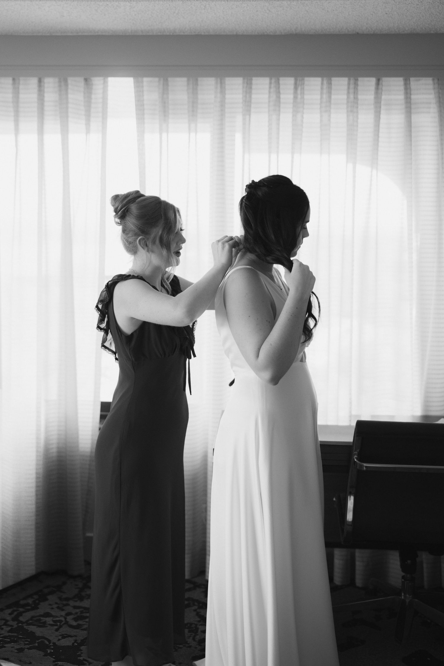 Bridesmaid helping bride button her white wedding gown in black and white photo by window.