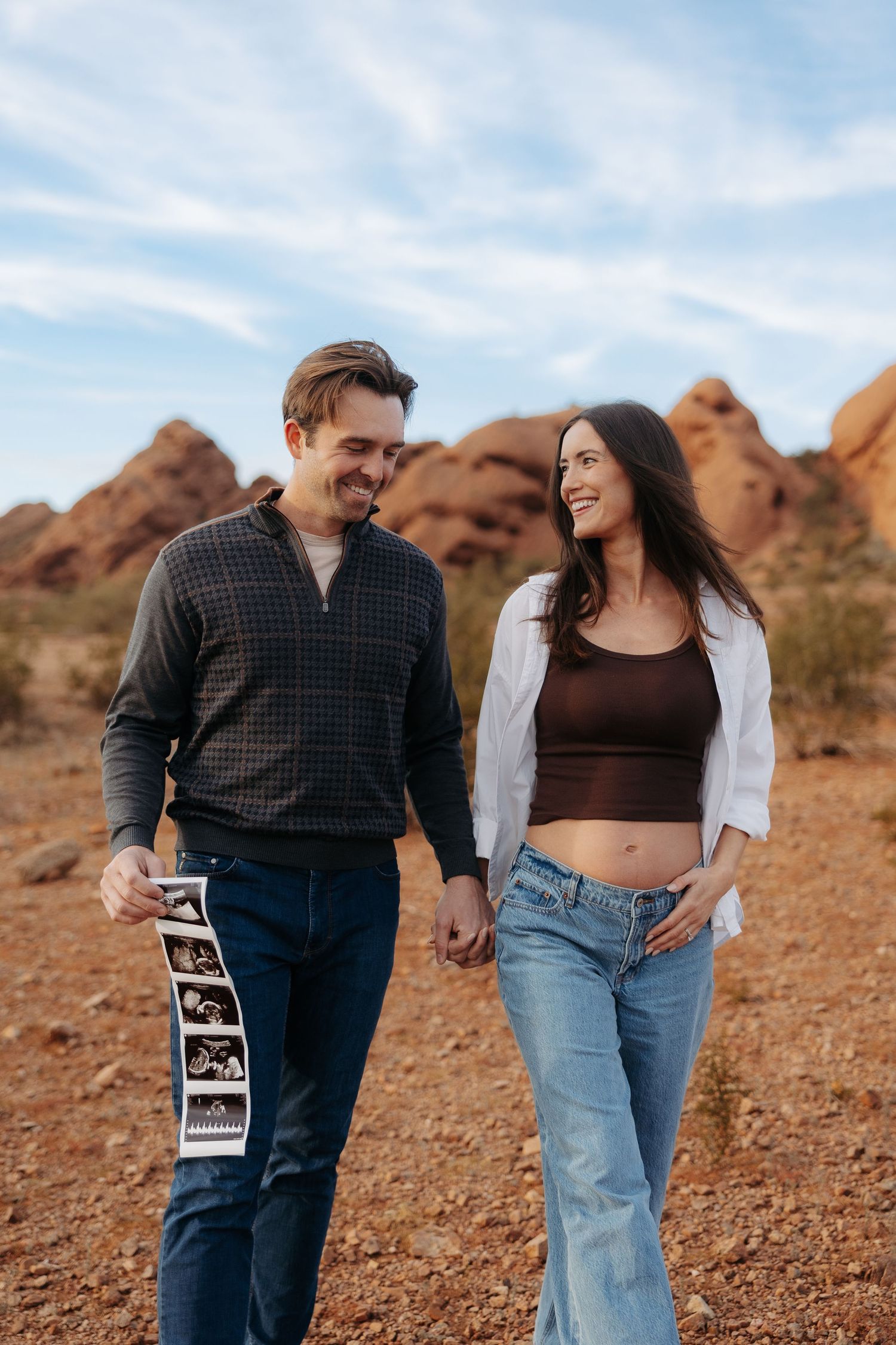 Smiling couple holding hands and ultrasound photos in a desert landscape with red rock formations, pregnancy announcement.