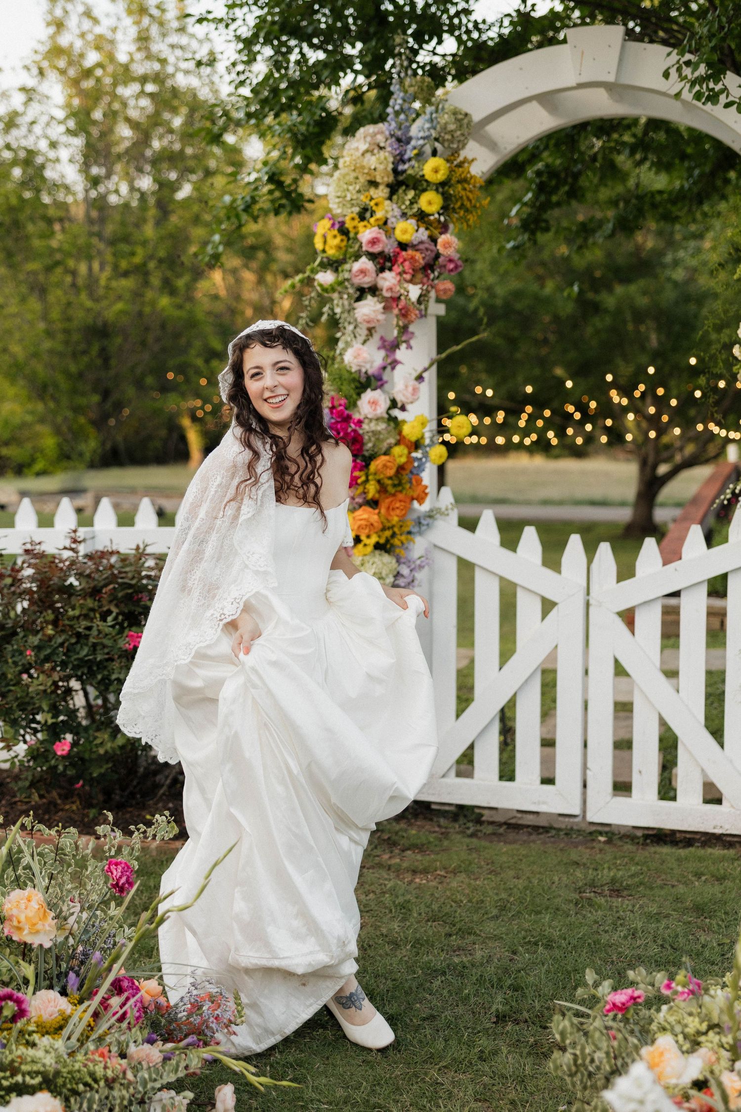 Smiling bride in white gown lifting dress near floral arch with colorful blooms and white picket fence.