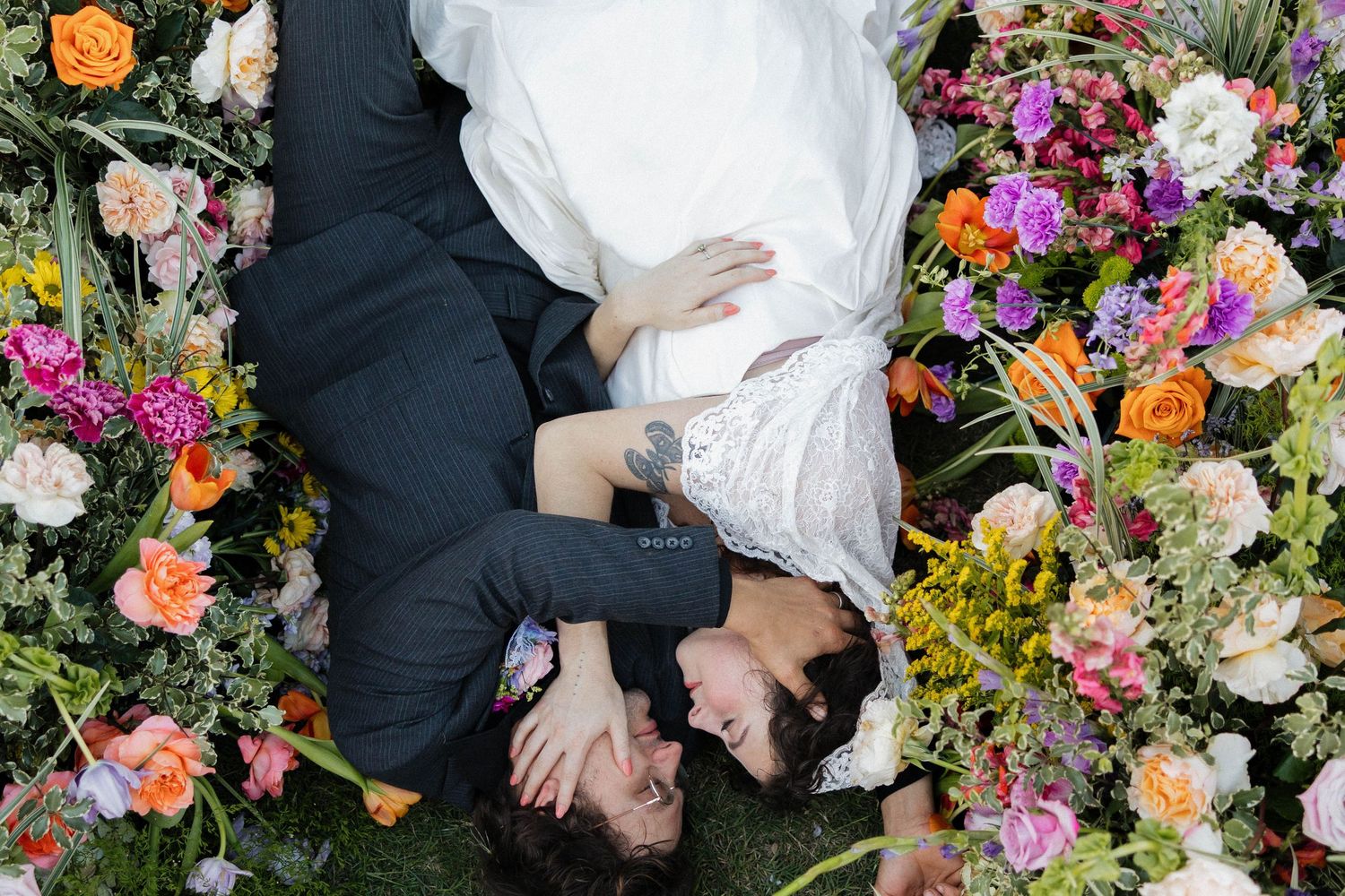 Aerial view of bride and groom lying among colorful wedding flowers, sharing a romantic moment surrounded by vibrant blooms.