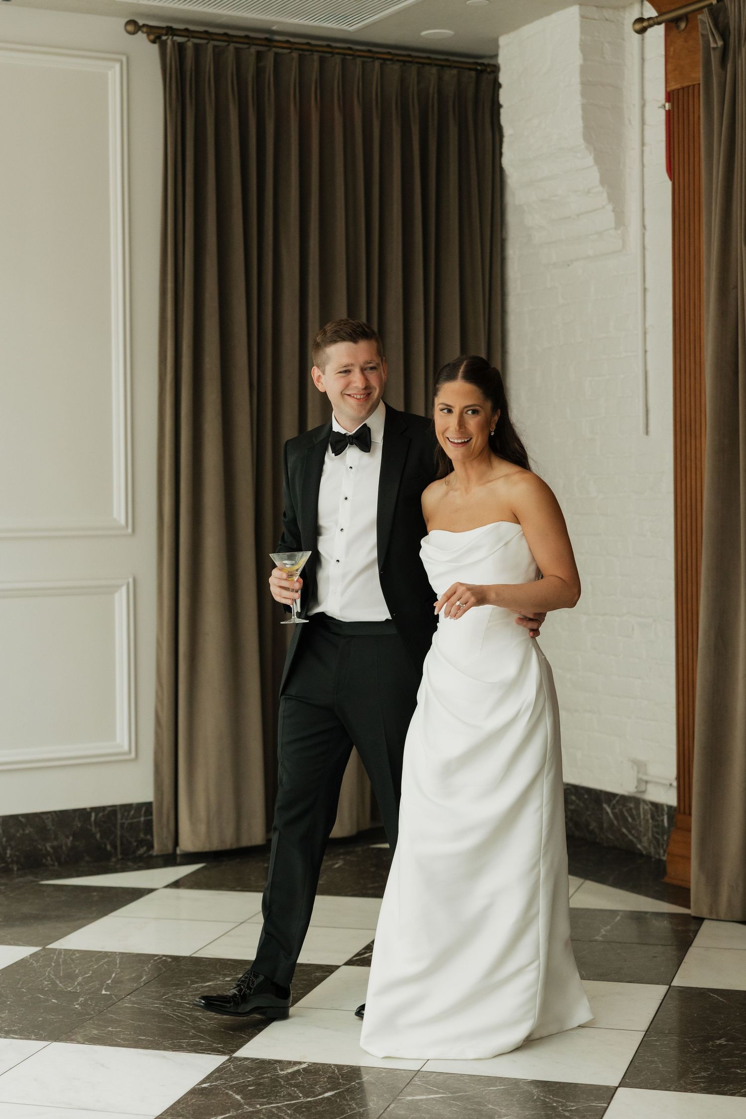Elegant bride in strapless white gown stands with tuxedo-clad groom holding cocktail on black and white checkered floor.