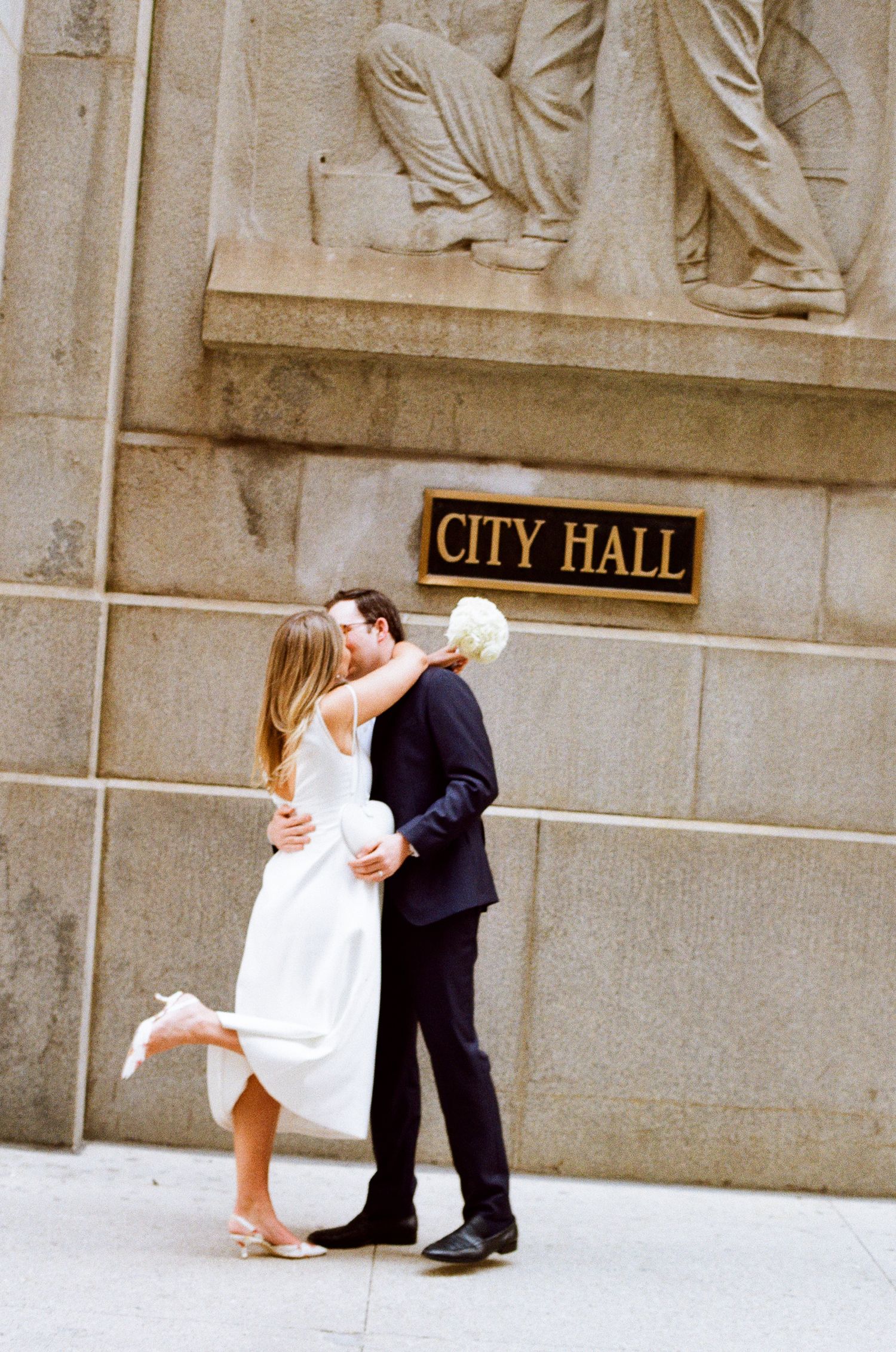Newlywed couple sharing a romantic kiss outside City Hall, bride in white dress holding bouquet, groom in dark suit.