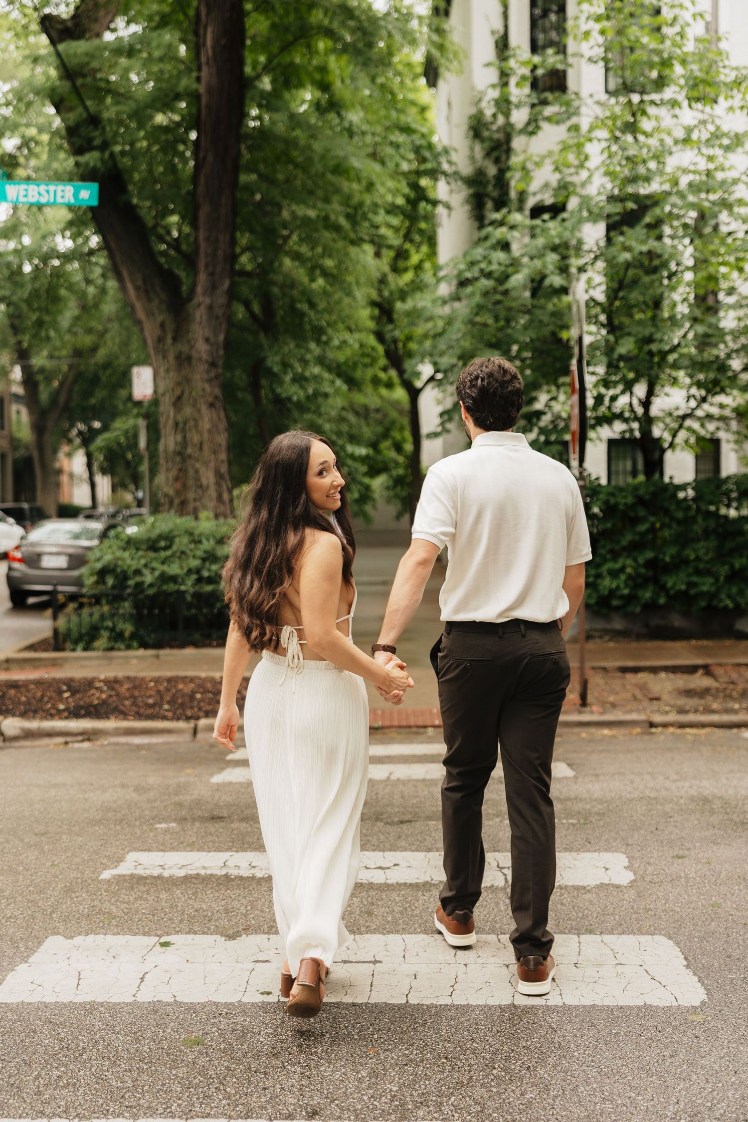 Couple holding hands crossing a zebra crossing on Webster Street, woman in white dress looking back, lush green trees lining the road.
