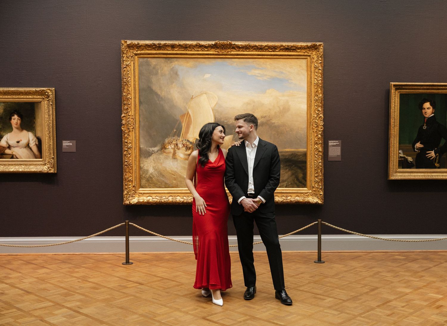 Two people in formal attire walk through an elegant art gallery with gold-framed paintings on display.