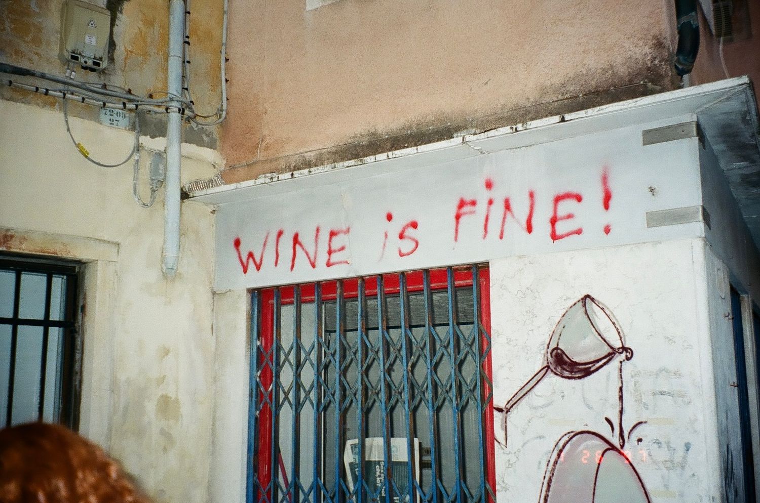 Graffiti on a wall reads 'WINE IS FINE!' with a simple red wine glass drawing next to metal bars on a window.