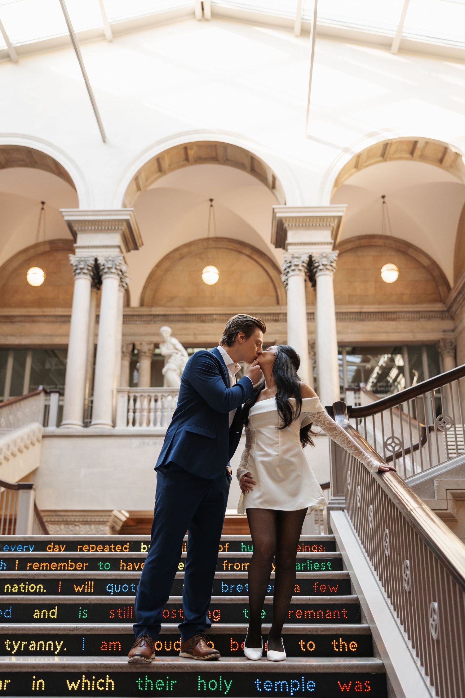 A couple shares a romantic moment on decorative steps inside a building with grand arched columns.