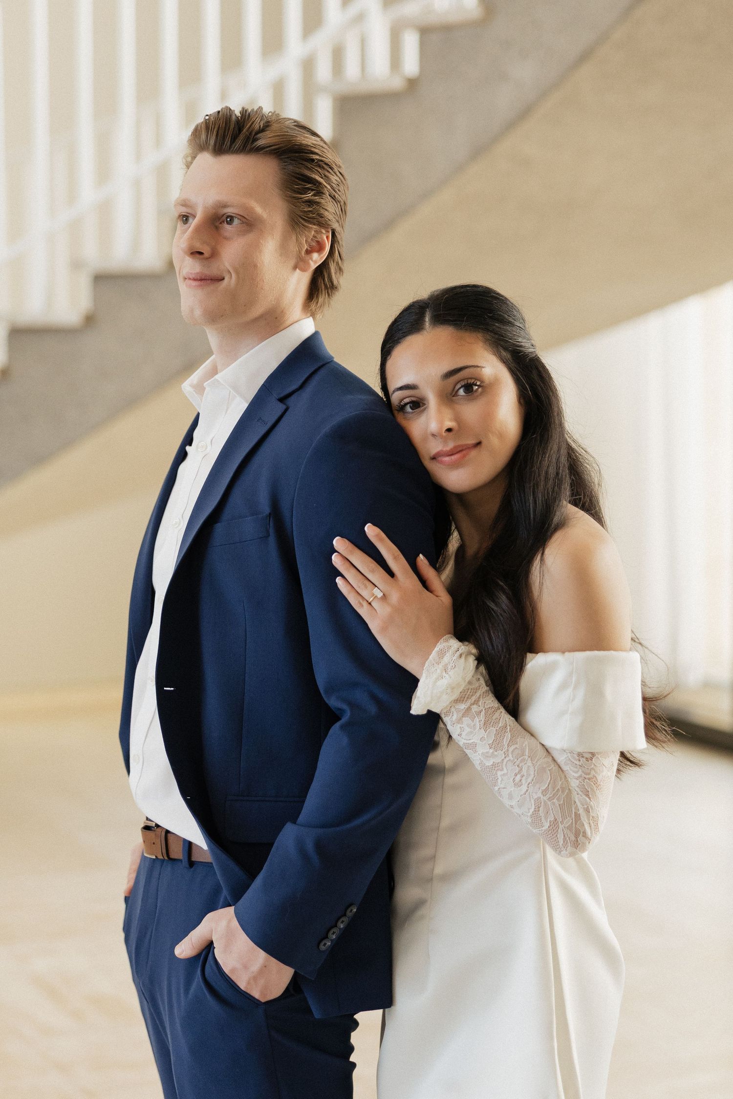 Couple embracing in front of white staircase in bright interior space.