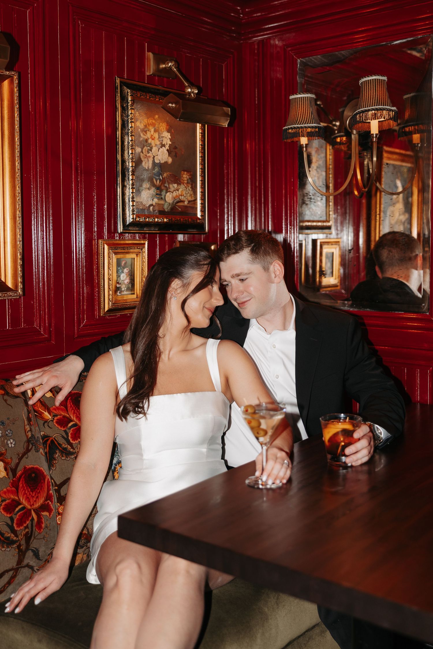 A romantic couple enjoying drinks together in a luxurious red-walled bar with ornate framed artwork.